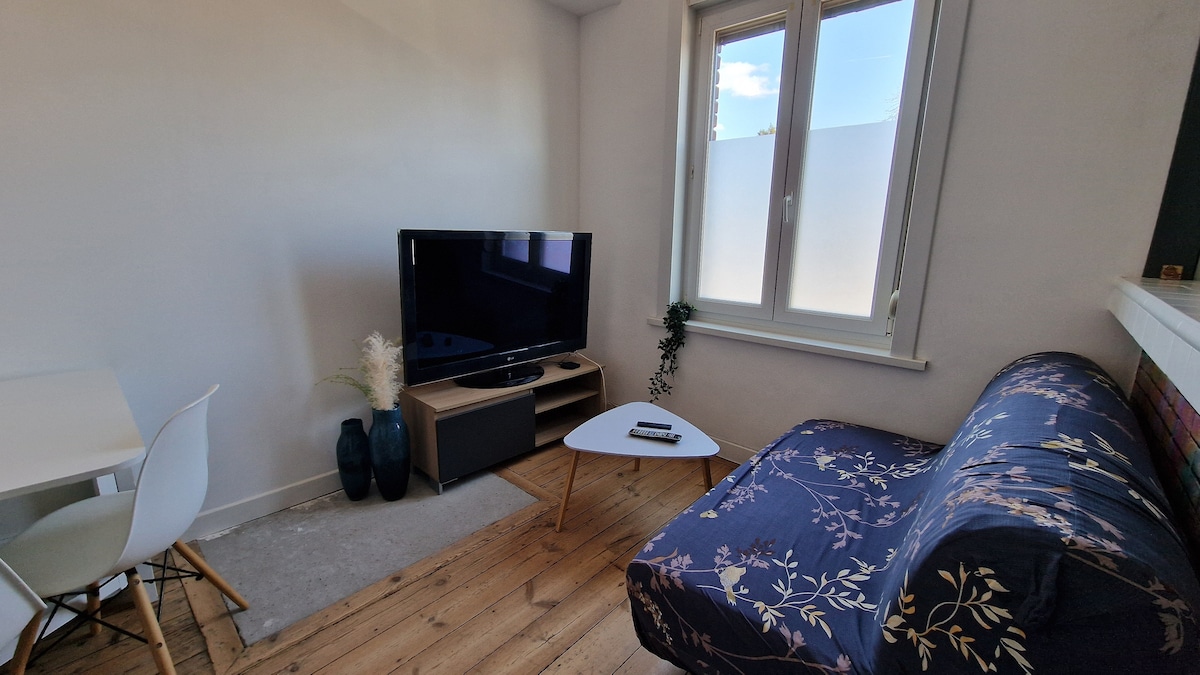 A comfortable seating area is depicted, featuring a sofa with a floral pattern and a small coffee table. A large television is positioned on a media unit, with a window allowing natural light to enhance the space. Two vases are placed on the media unit, adding a touch of decor.