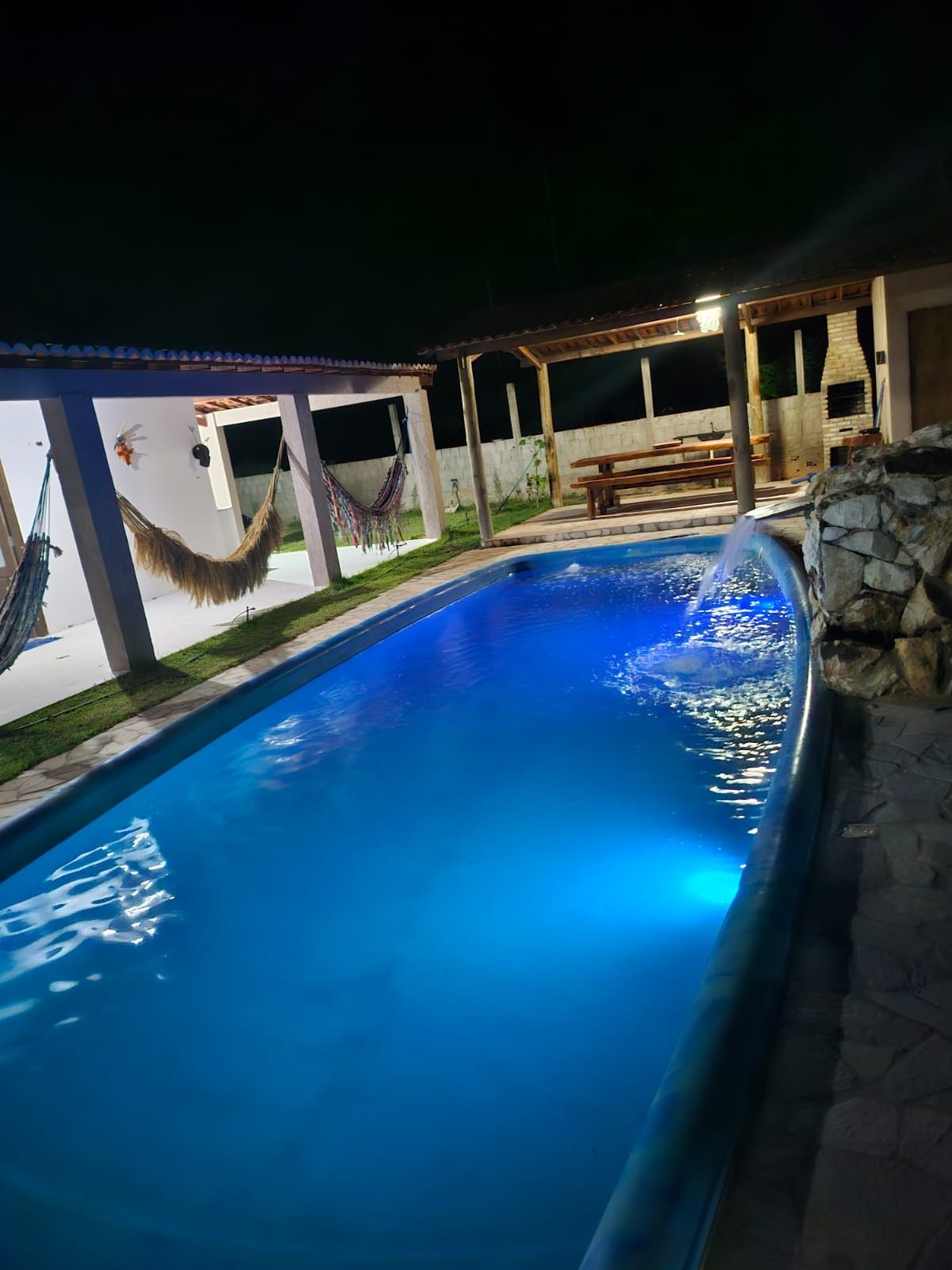 A tranquil pool area is illuminated in soft blue light, surrounded by stone accents. Comfortable hammocks hang nearby, providing a relaxed seating option. An outdoor dining table is visible in the background, situated under a sheltered area that enhances the serene ambiance.