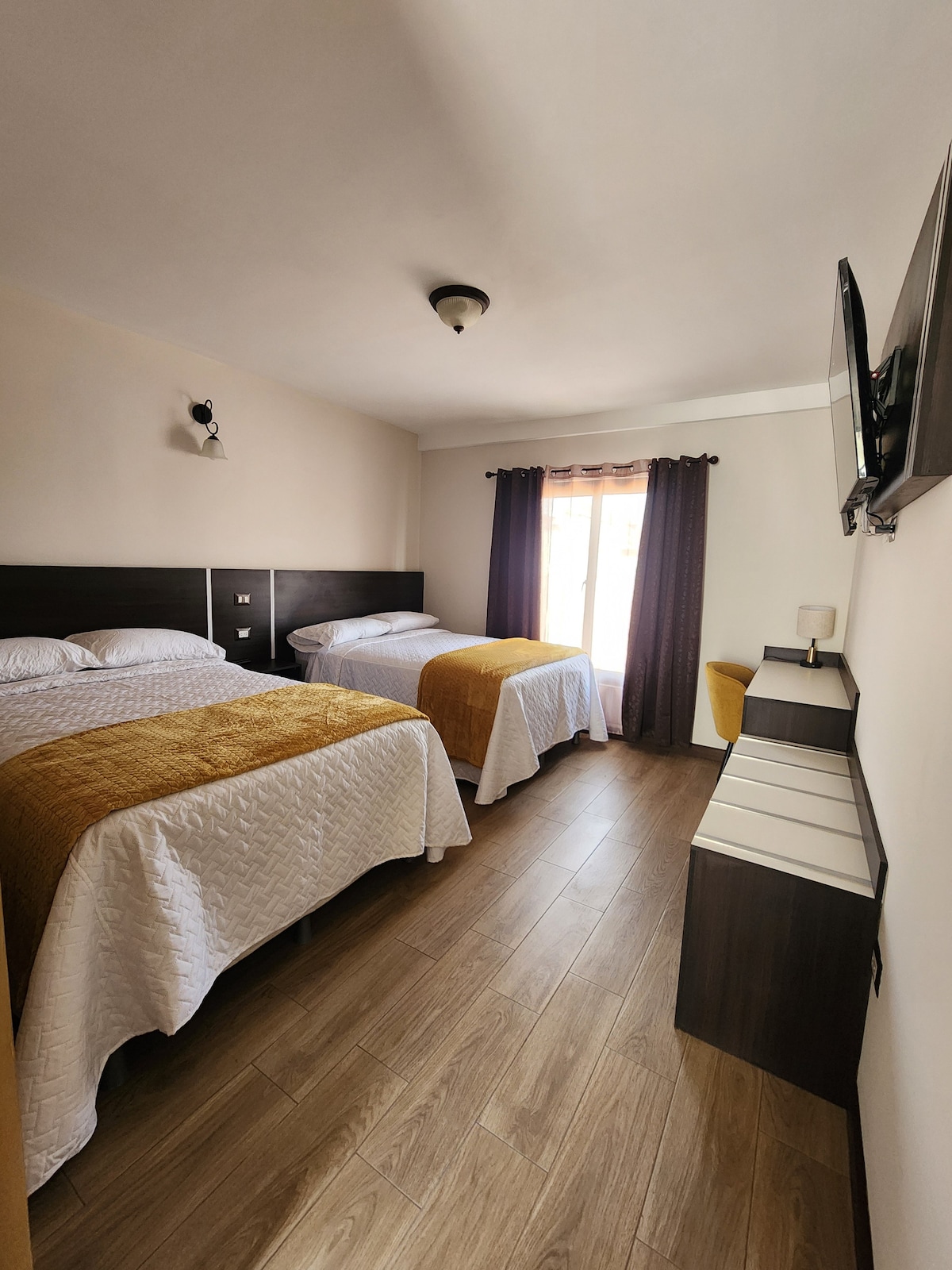 A bright and airy guest room features two spacious beds with elegant white bedding complemented by golden throws. Natural light enters through a window adorned with flowing dark curtains, while a small wardrobe and a television add functional elements to the space.