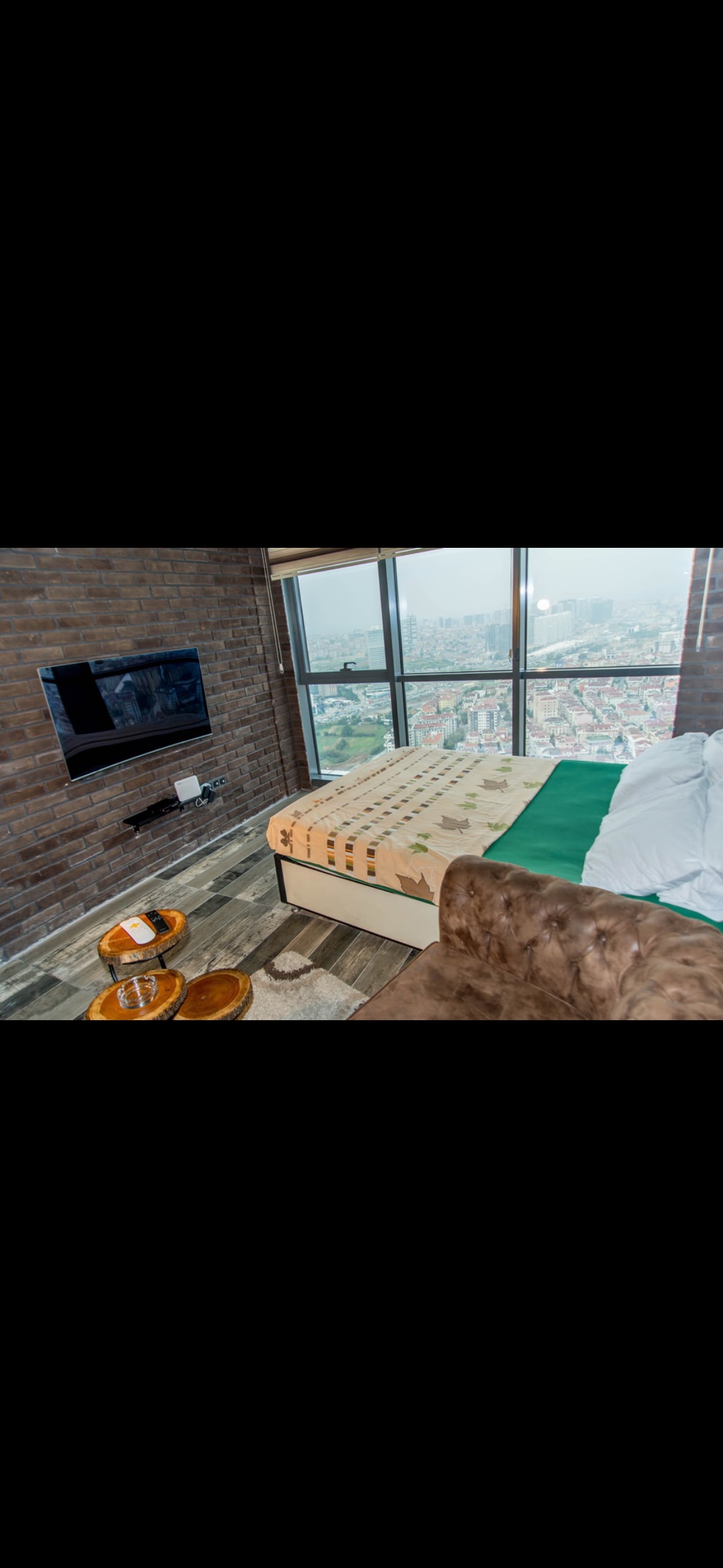 A cozy corner features a spacious bed with a green blanket against a backdrop of exposed brick walls. A flat-screen TV is mounted nearby, and large windows offer a panoramic view of the city skyline. Stylish wooden tables sit beside a comfortable seating area.