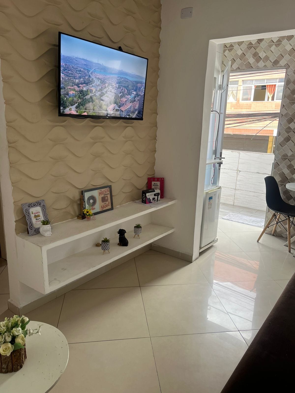 A cozy living area features a mounted flat-screen TV above a white shelf. Decorative items and a small floral arrangement are placed on the shelf. A doorway leads to a balcony, allowing natural light to fill the space.