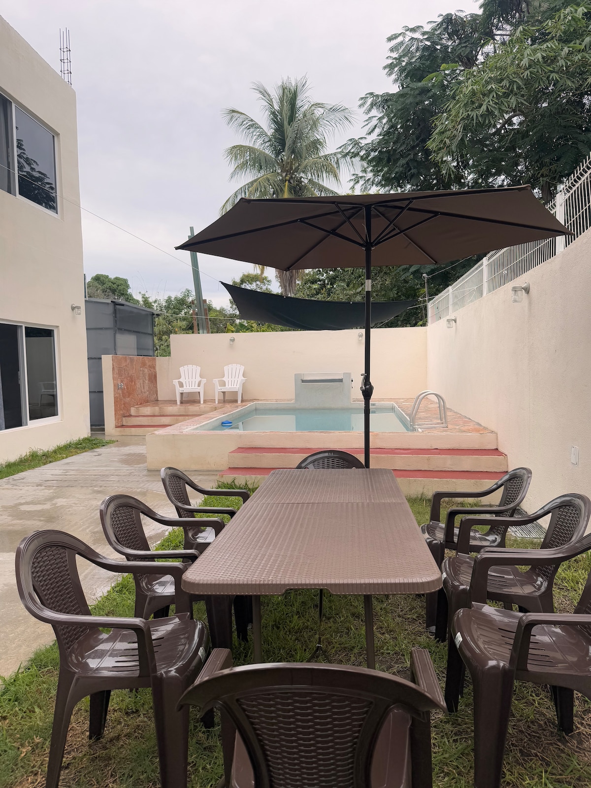 An outdoor dining area is set beneath a shaded umbrella, featuring a large table surrounded by plastic chairs. A small private pool is visible in the background, flanked by two lounge chairs, all amidst a grassy space.