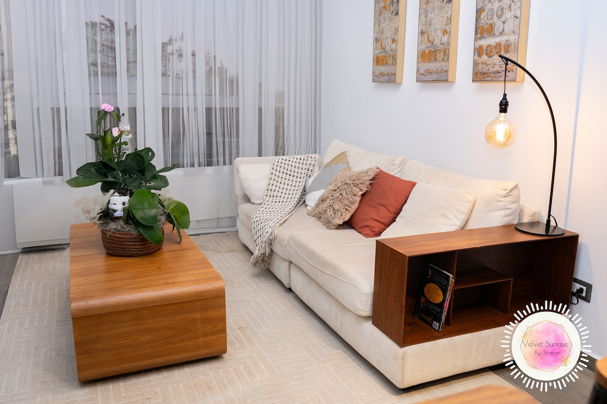 A light-filled living area features a cozy, L-shaped couch adorned with a mix of cushions. A wooden coffee table with a plant centerpiece adds a touch of nature, while illuminated by a stylish pendant light. Sheer curtains allow natural light to flow in softly.