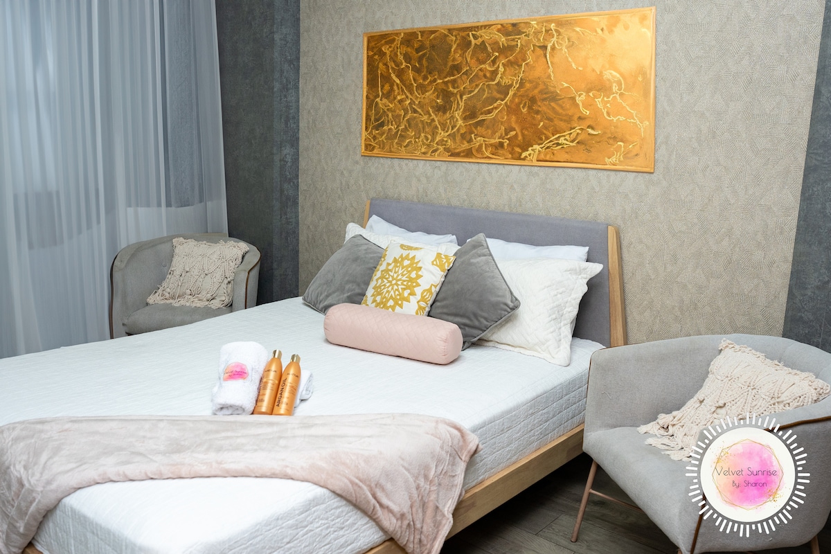 A cozy bedroom is showcased with a spacious bed adorned with soft linens and decorative pillows. Two armchairs sit beside the bed, complementing the elegant wall art above. Natural light filters through sheer curtains, highlighting the warm tones of the room.