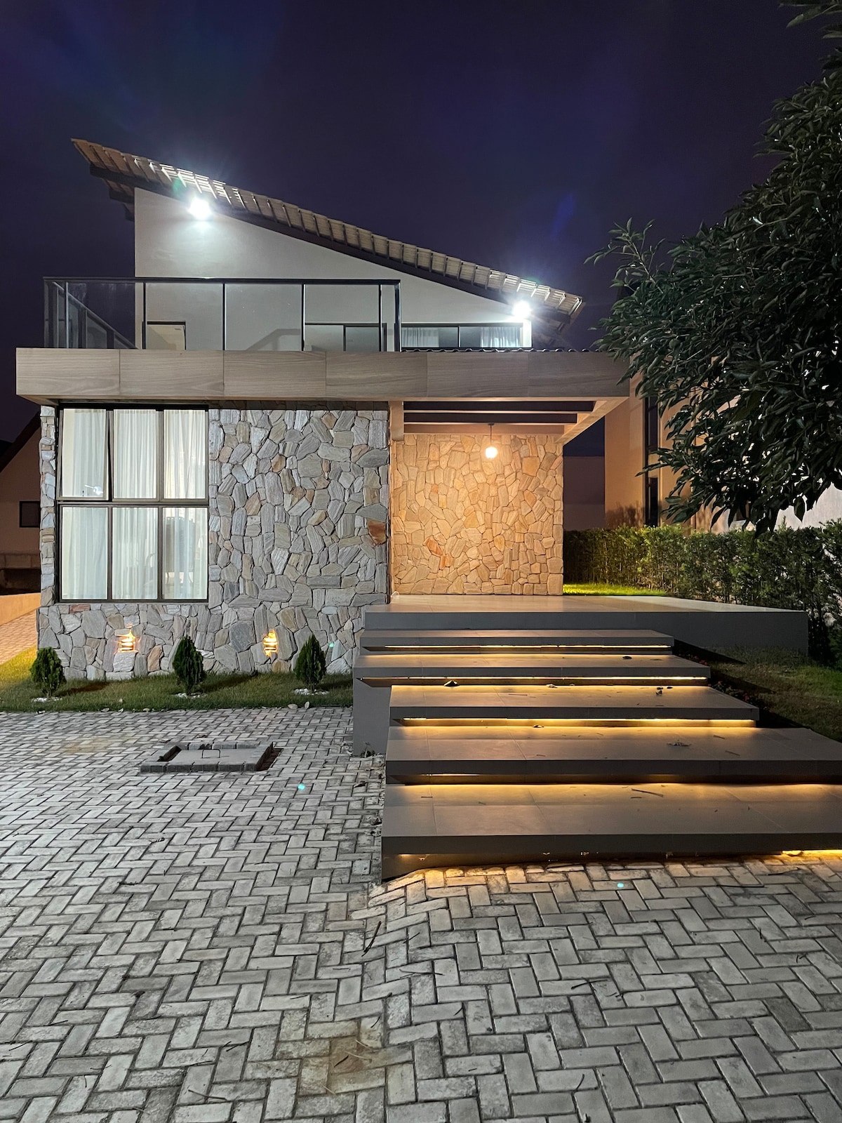 The exterior of the house features a modern design with a stone facade and wooden accents. Soft lighting illuminates the entry steps leading to the porch. Lush greenery surrounds the property, creating an inviting and well-maintained atmosphere.