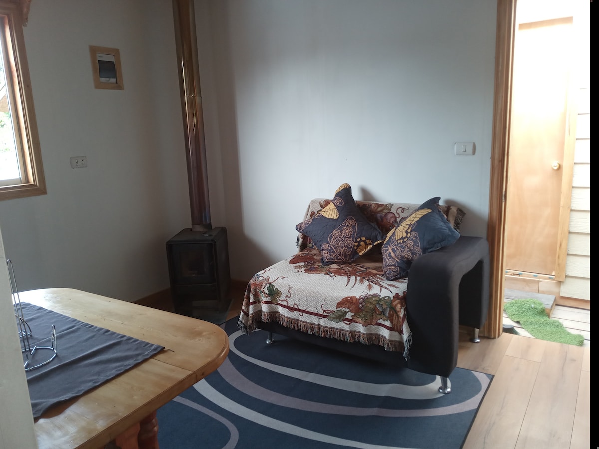 A cozy seating area features a dark sofa adorned with patterned cushions, positioned near a small wood stove. Natural light enters through a window, illuminating the space. A wooden table rests nearby, with a simple place setting on a textured tablecloth.