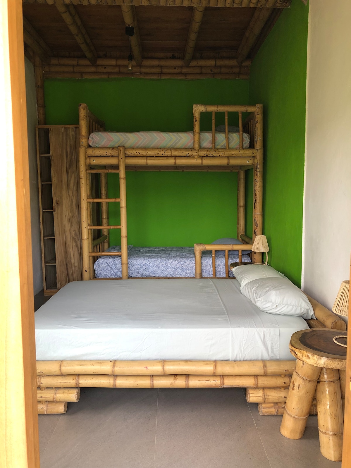 A bamboo-furnished sleeping area features a spacious bed and a bunk bed against a bright green wall. Two bedside lamps are placed next to the beds, and a wooden shelving unit is positioned nearby, completing the cozy environment.