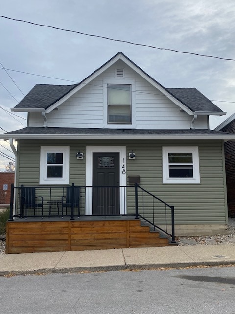 A two-story house features a combination of light green and white siding. The front entrance, highlighted by a dark door, is complemented by two windows on either side. A small wooden porch with black railings provides access, while outdoor seating is visible near the entrance.