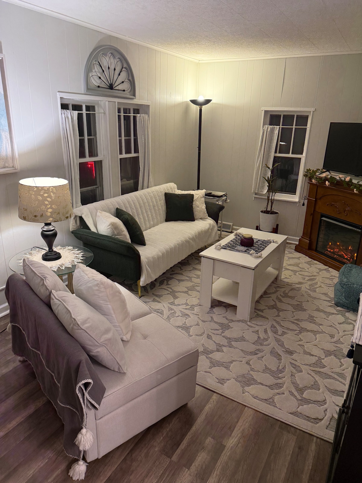 A cozy living area is depicted, featuring a light-colored sofa and a dark green armchair. A small, rectangular coffee table is centered, with a decorative item placed atop. Soft lighting is provided by a standing lamp, and the warm glow of an electric fireplace is visible along the wall.
