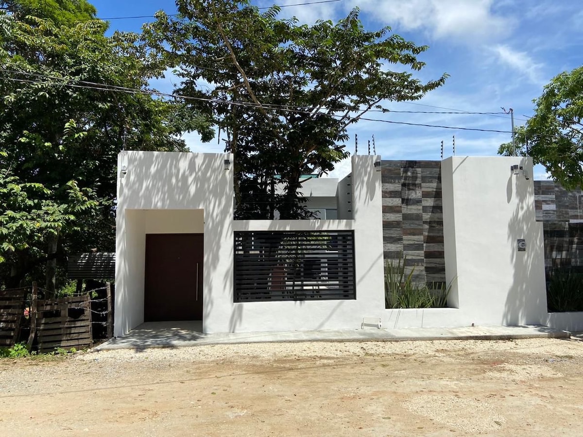 The exterior of the property features a modern facade with clean lines and a combination of white walls and natural stone accents. A wooden front door is positioned to the left, while horizontal slats provide privacy for a window. Lush greenery surrounds the entrance.