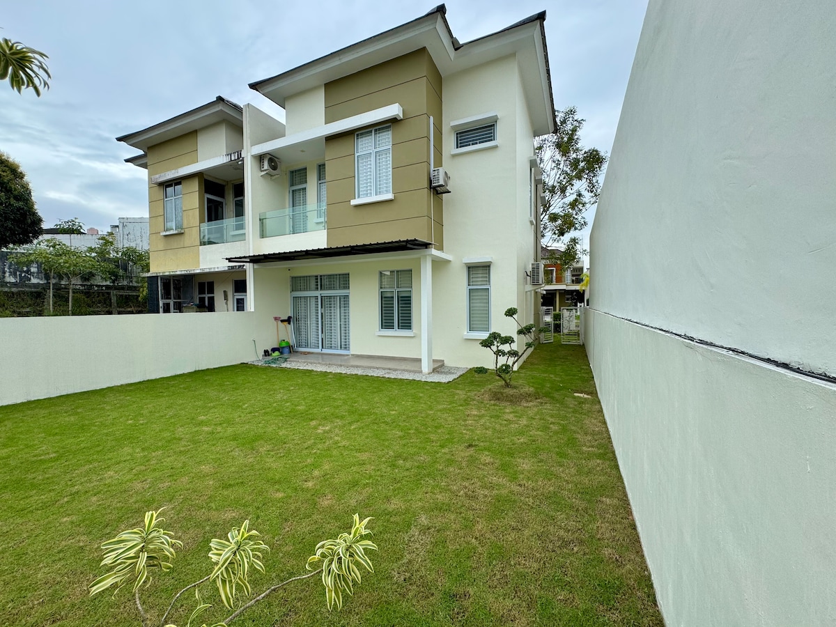 A spacious yard is surrounded by a high wall, featuring well-maintained green grass. The two-story house is visible, showcasing large windows and a balcony. A small garden area is present, complemented by landscaping elements.