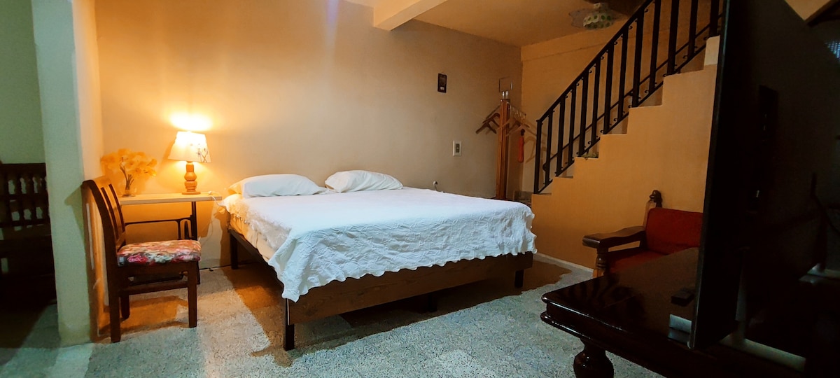 A well-lit sleeping area features a spacious bed covered with a light-colored blanket. A small table with a lamp is placed beside the bed. A wooden chair adds to the decor, while a staircase is visible in the background, leading to the upper level.