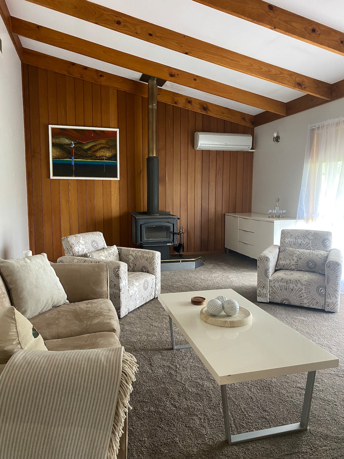 A cozy living area features wooden walls with a high, beamed ceiling. Comfortable seating, including two armchairs and a sofa, surrounds a coffee table. A wood-burning stove is positioned against the wall, while natural light enters through a nearby window, complemented by light curtains.