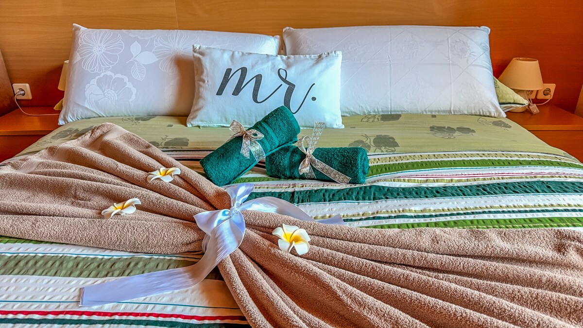 A cozy bedroom setting features a spacious bed with decorative pillows and a soft blanket. Towels, tied with ribbon, are positioned neatly alongside soft floral accents, creating a serene and welcoming atmosphere.