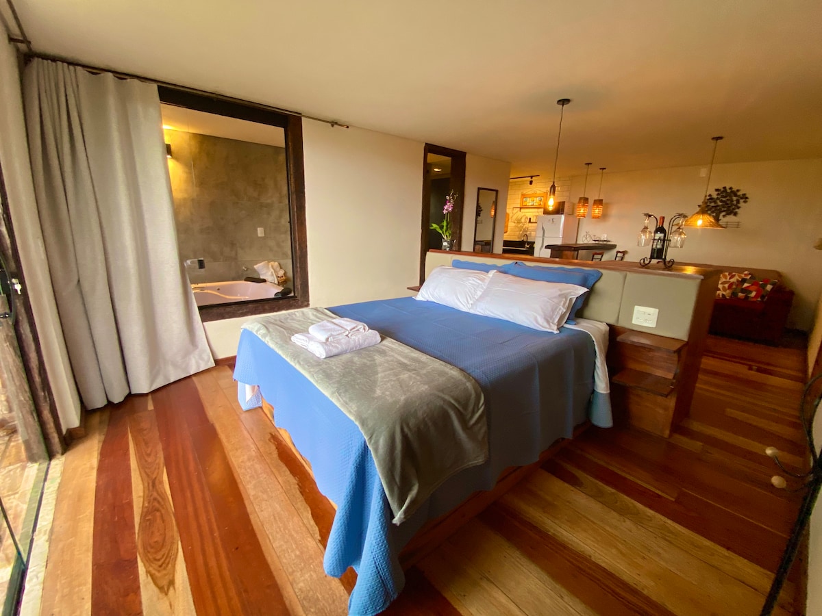 The spacious bedroom features a comfortable bed with a blue cover and layered blankets. Wooden floors are prominent, with natural light flowing in through large windows. A separate living area can be glimpsed through an adjoining doorway, enhancing the open feel of the loft.