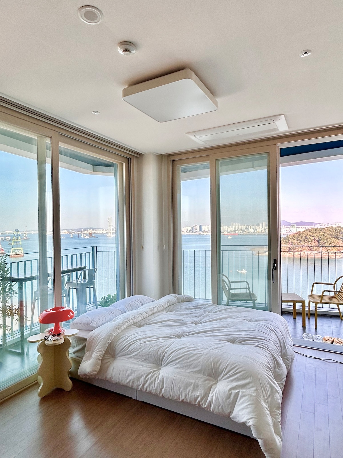 A spacious sleeping area features a large bed covered with a white comforter. Floor-to-ceiling windows provide unobstructed views of the ocean and surrounding landscape. A small table and two chairs are visible on the balcony, creating an inviting outdoor space.