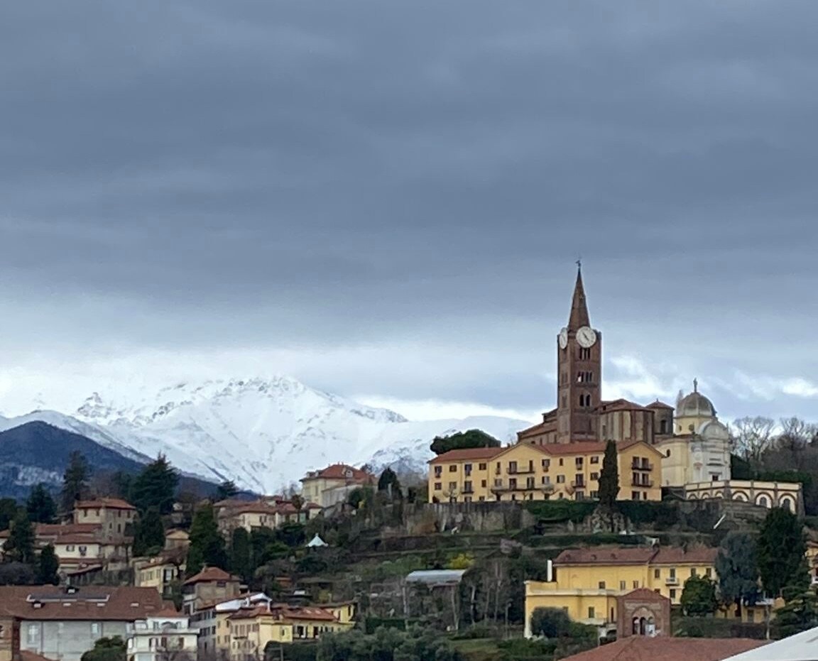 A scenic view features a historic clock tower and church amidst a charming town set against a backdrop of snow-capped mountains. The landscape is characterized by a mix of colorful buildings and lush greenery, reflecting a tranquil atmosphere under a cloudy sky.