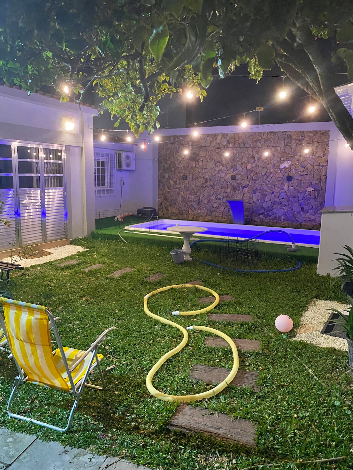 A private backyard features a heated pool illuminated with soft blue lighting, surrounded by a grassy area. String lights hang above, adding a gentle glow. A yellow and white striped lounge chair sits on a pathway, leading to the adjacent structures.
