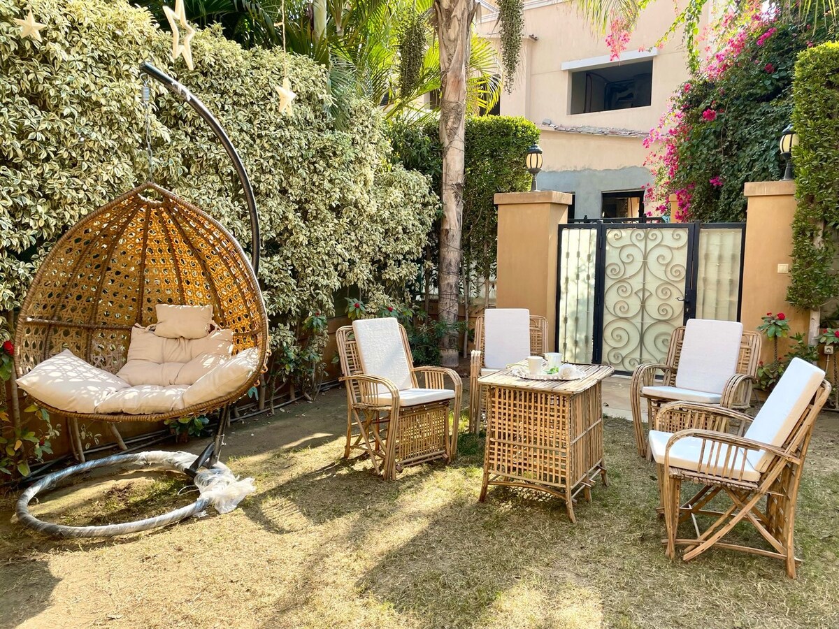 An outdoor seating area is presented with a cozy hanging chair and four wicker chairs arranged around a small table. Lush greenery and blooming flowers create a private garden space, complemented by decorative fencing that provides a serene atmosphere.