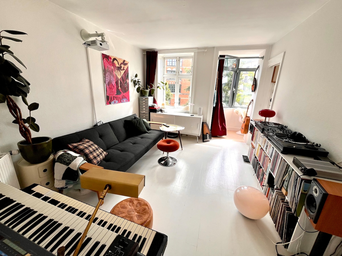A bright living room is highlighted by a black couch and a round coffee table. A vibrant artwork decorates one wall, while a collection of vinyl records is displayed on the opposite side. Natural light enters through large windows, complemented by red drapes.