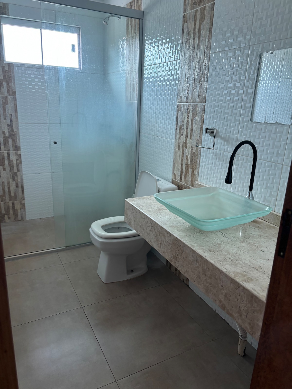 A modern bathroom features a glass shower enclosure with textured tiles. A sleek, transparent sink basin rests on a stone countertop, complemented by a minimalist faucet. Natural light enters through a window, highlighting the clean lines and contemporary design.