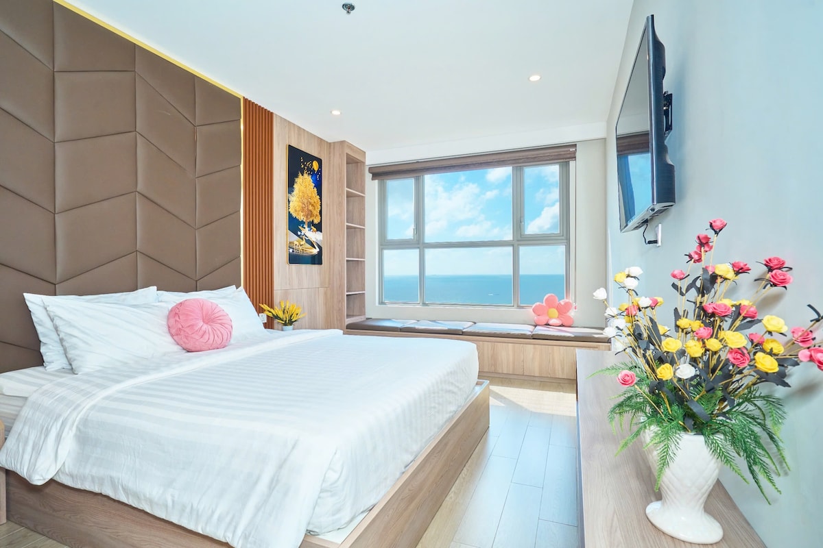 A spacious sleeping area features a large bed with crisp white linens and decorative pillows. A well-lit window offers a view of the sea, while a flower arrangement adds a touch of color to the room.