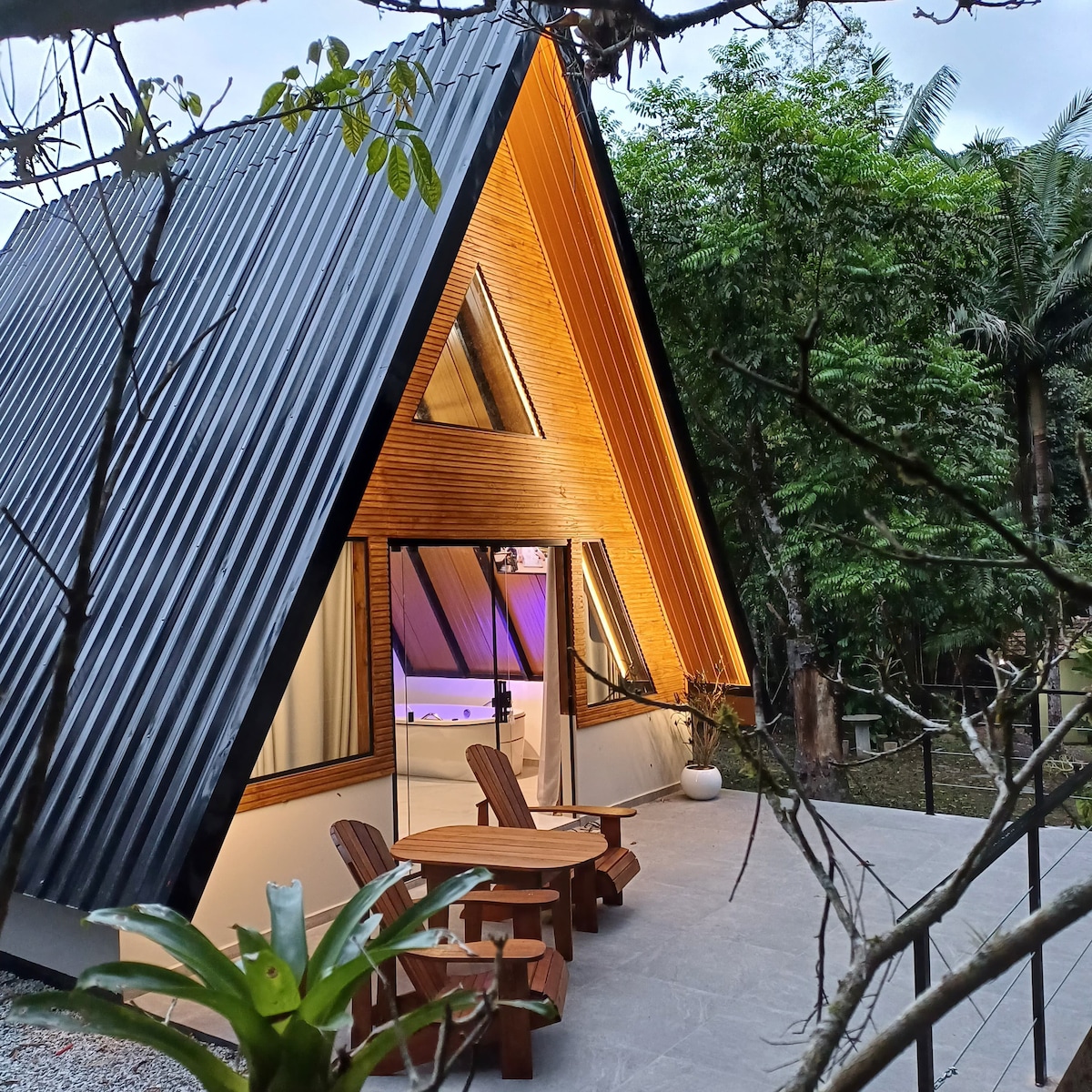 A modern A-frame chalet is depicted with a striking wooden facade and expansive glass doors. The outdoor area features a small seating arrangement with two wooden chairs and a table. Lush greenery surrounds the structure, enhancing the tranquil setting.