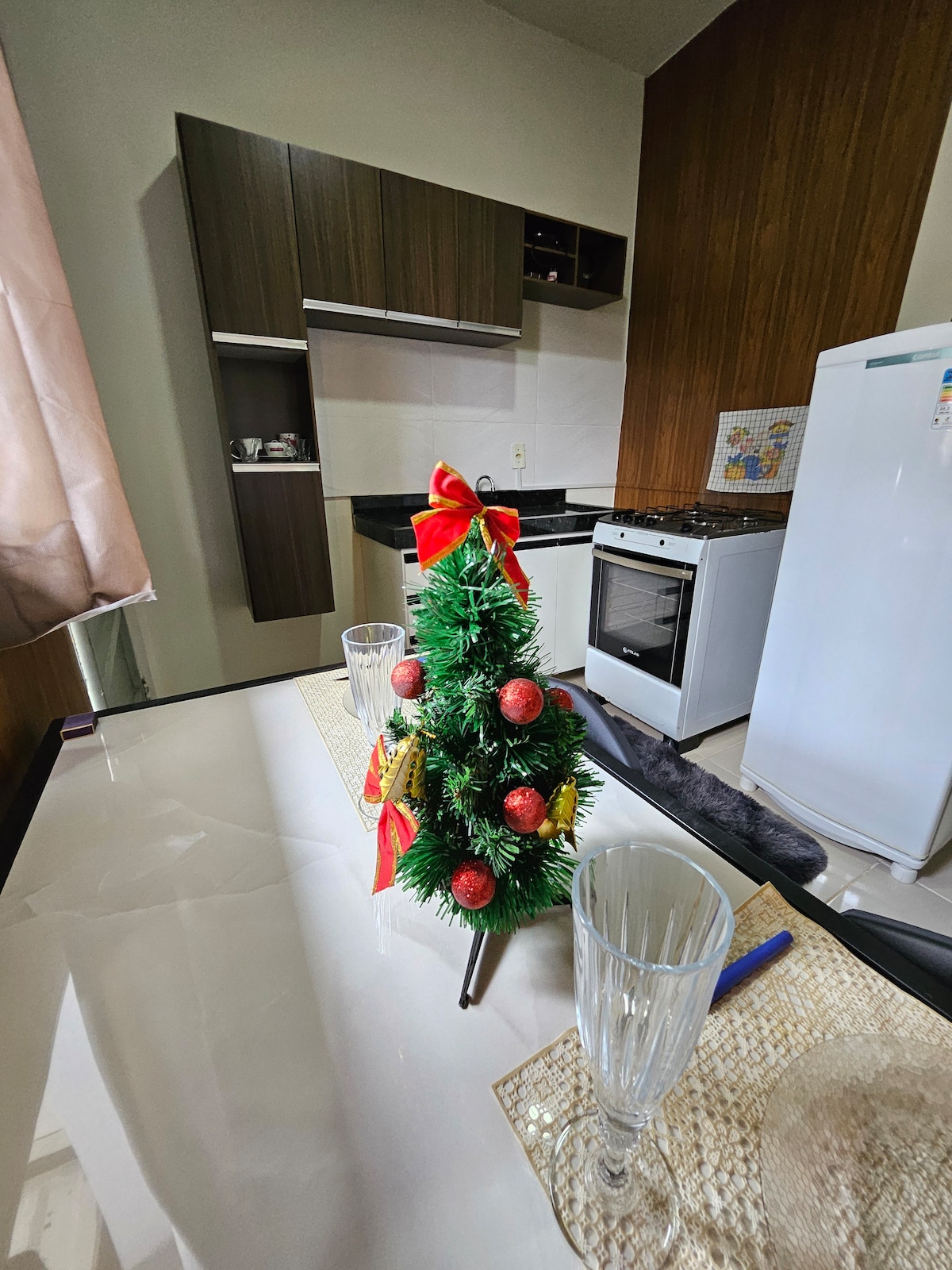 A modern kitchen area features dark wood cabinetry and a white countertop. A small, decorative Christmas tree with red ornaments is centered on the dining table, which is set with clear glassware. A white refrigerator and a stove are positioned in the background.