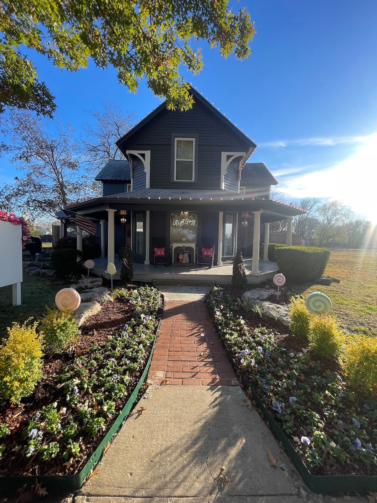 A charming historical home is presented with a dark exterior and a welcoming front porch. Colorful plants and a brick pathway lead to the entrance, framed by decorative garden elements. Sunlight creates a warm glow, enhancing the inviting appearance of the lush landscaping.