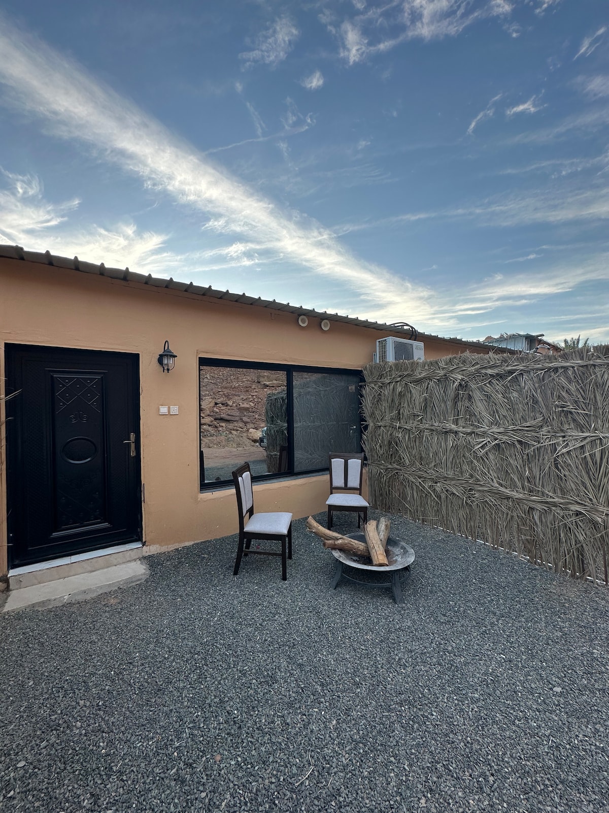 An outdoor seating area is presented with two chairs positioned around a fire pit. A rustic privacy wall made of natural materials stands alongside the property, while a clear sky with wispy clouds creates a serene backdrop.