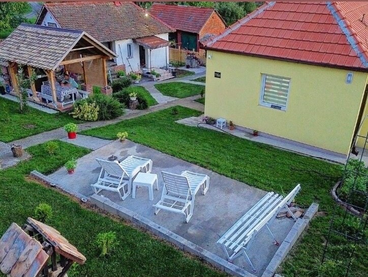 An overview of the outdoor space reveals a sunbathing area with lounge chairs on a paved surface. Surrounding gardens feature well-maintained grass and decorative plants. A shaded pavilion offers seating and is positioned nearby, contributing to a relaxed atmosphere in the countryside setting.