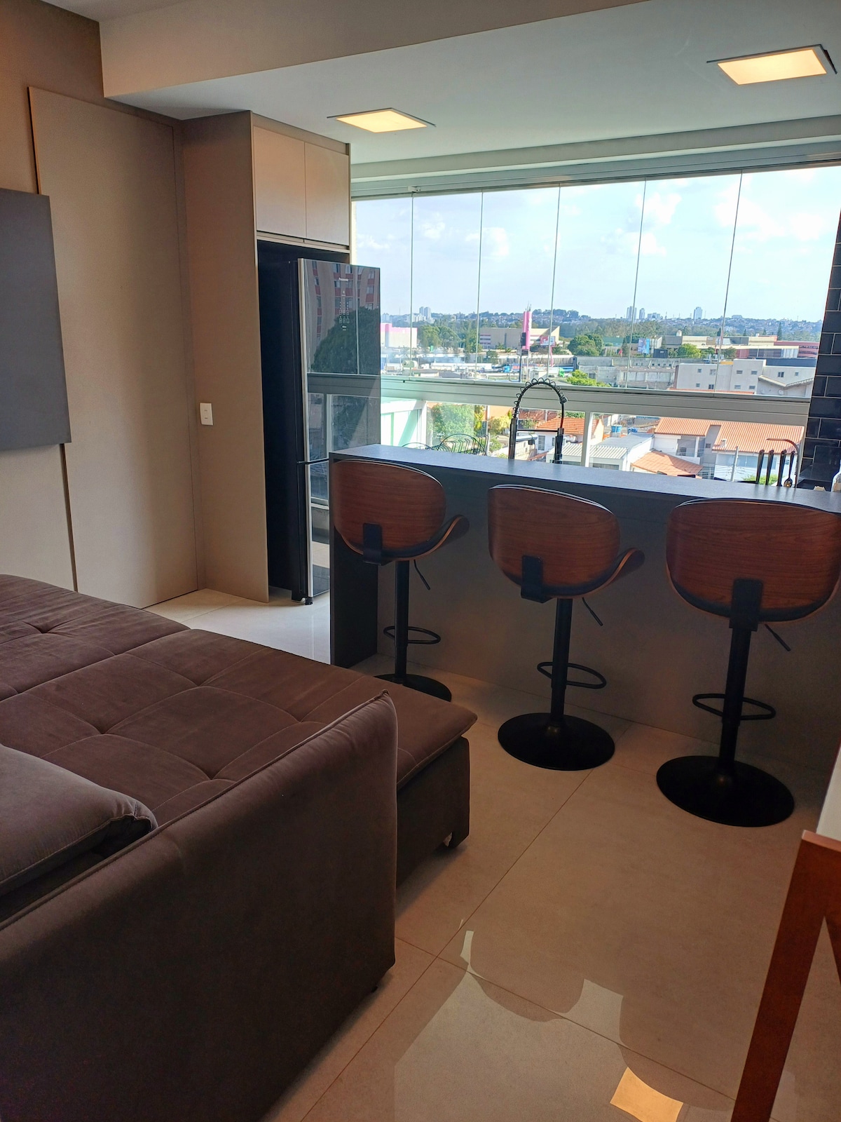 A modern studio space features a cozy seating area and a kitchen bar with three stylish stools. Large windows provide a view of the cityscape, allowing natural light to brighten the room. Neutral tones create a relaxed atmosphere.