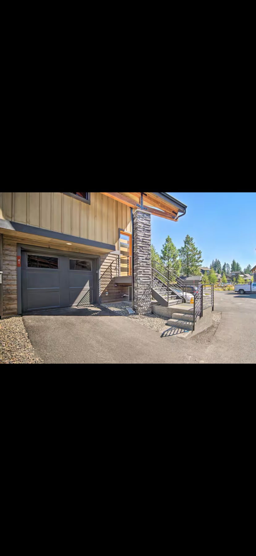The exterior view showcases a two-story building with a stone-accented support column. A garage with a sleek door is visible, along with a pedestrian pathway leading up to the entrance. Surrounding trees add a natural touch to the setting.