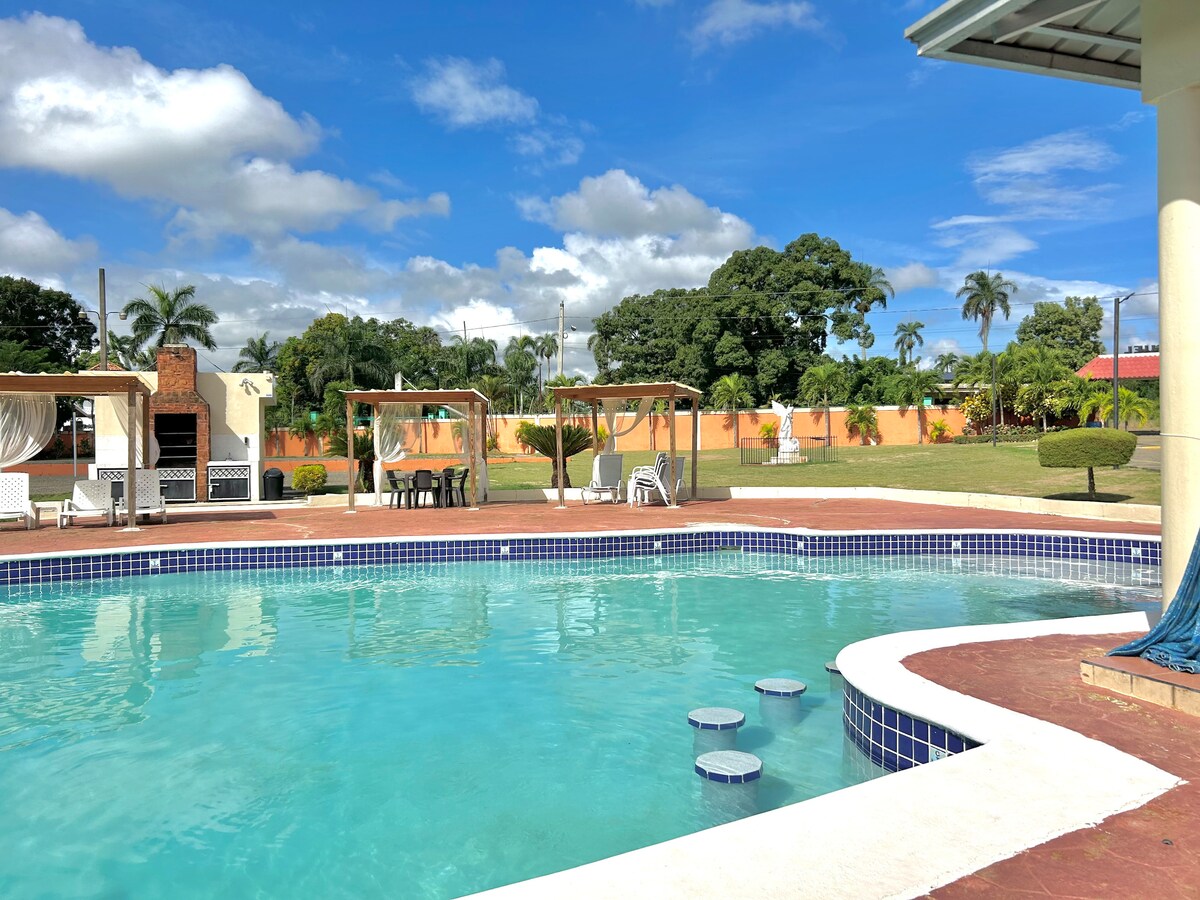 A refreshing swimming pool is featured with a gentle curve, surrounded by a spacious patio. Lounge areas with shade structures are visible, along with greenery and palm trees in the background. Bright blue skies and soft clouds create a serene setting.