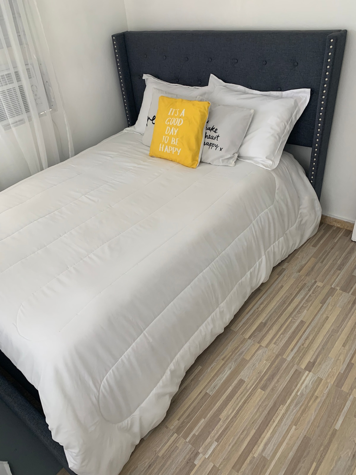 A spacious bed is dressed in crisp white linens, complemented by decorative pillows featuring motivational phrases. A bright yellow accent pillow adds a pop of color. Natural light filters through sheer curtains, casting a gentle glow on the warm wooden flooring.
