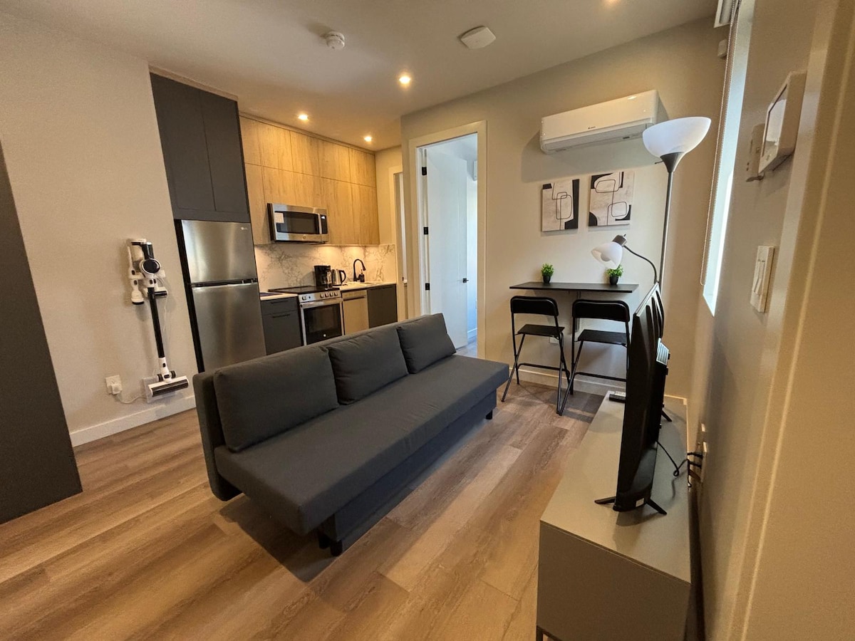 A modern living area is showcased with a grey sofa positioned centrally, complemented by a dining table with two black chairs. The kitchenette features sleek cabinetry and stainless steel appliances. Natural light filters through a window, illuminating the light wood flooring.