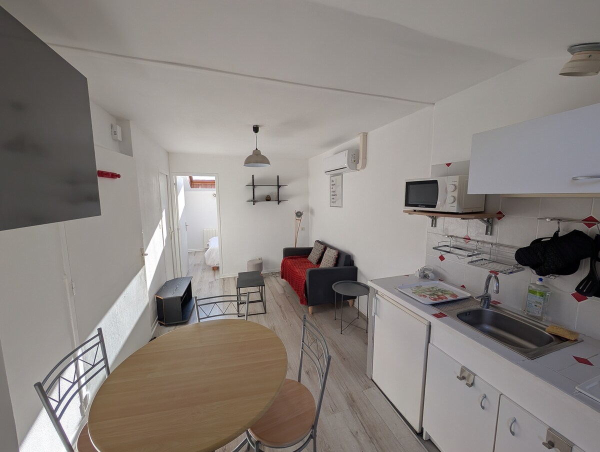 A functional studio space is presented, featuring a small dining table surrounded by metallic chairs. A seating area with a dark sofa and red accents is visible, alongside a compact kitchen equipped with essential appliances. Bright light enters through large windows, enhancing the airy atmosphere.