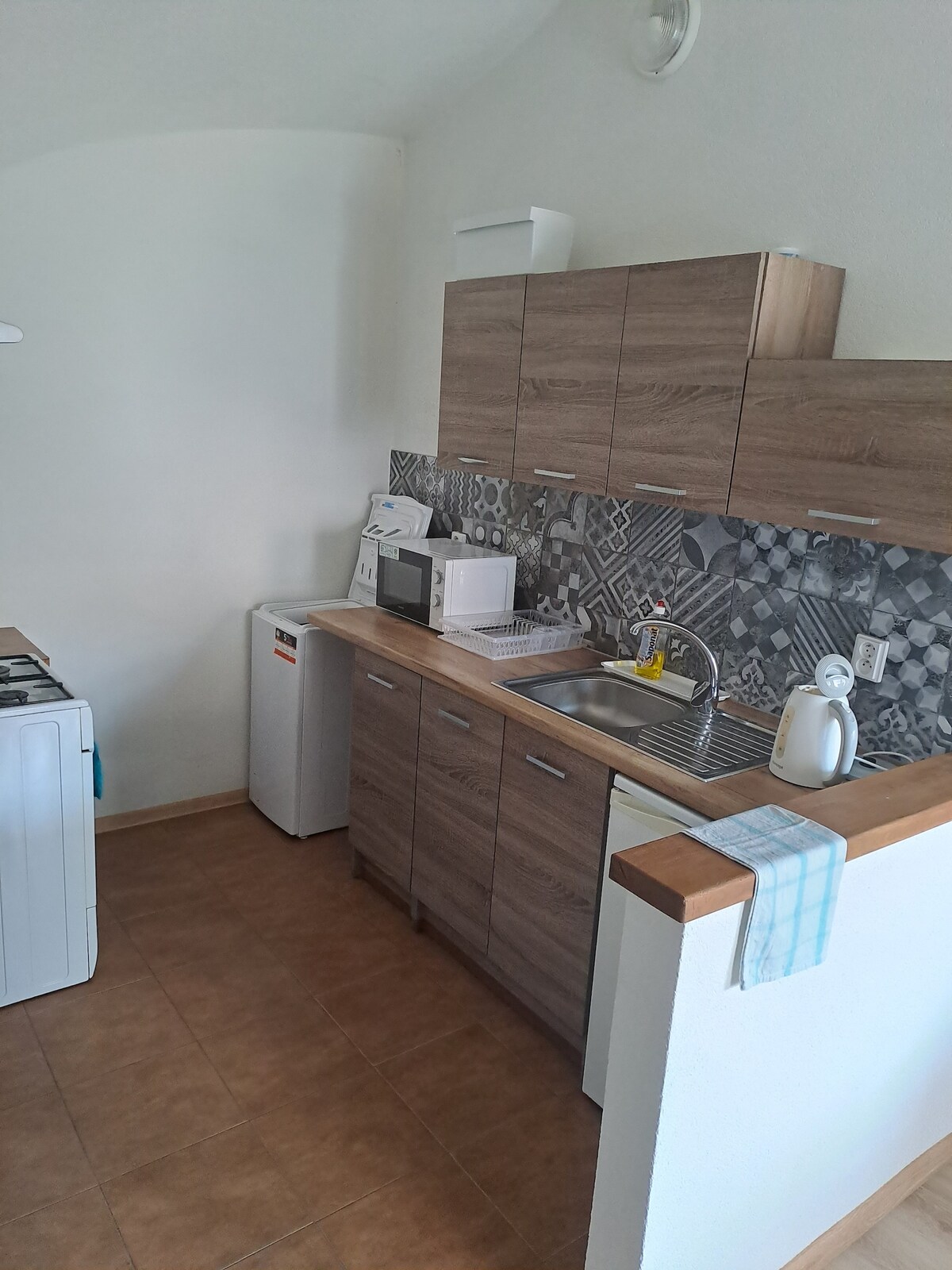 The kitchen features wooden cabinetry contrasted by a patterned backsplash. Essential appliances include a refrigerator, microwave, and sink, with a stove positioned nearby. A towel hangs neatly on the counter, and the space is lit by natural light from the nearby window.