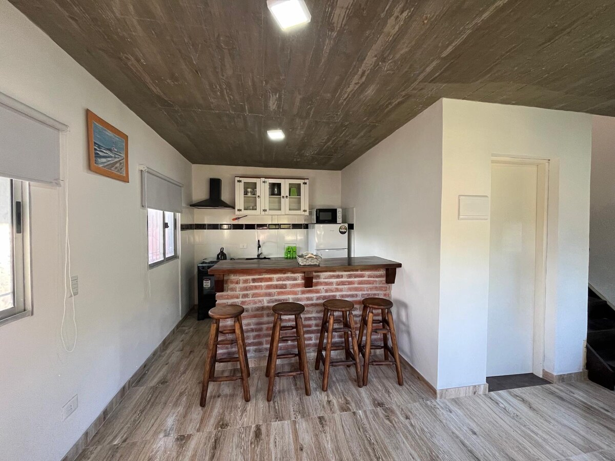 An inviting kitchen area features a wooden bar with three high stools. A brick accent wall complements the white cabinetry, equipped with essential appliances. Large windows provide natural light, illuminating the space with a bright and airy feel.