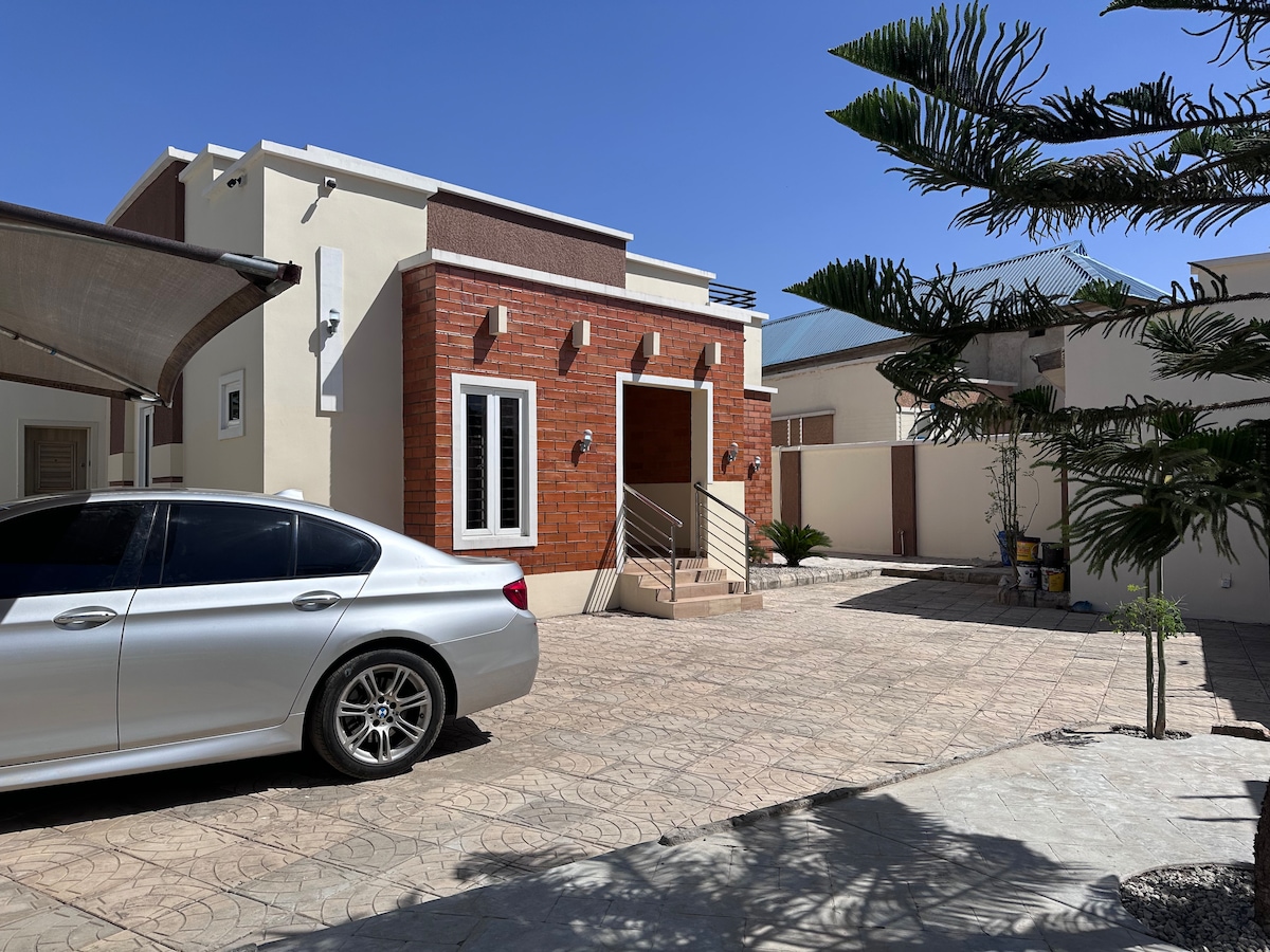 A modern house is showcased with a brick facade and a welcoming front entrance. A well-maintained driveway accommodates a silver car, with palm trees and landscaped plants adding a touch of greenery in the foreground against a clear blue sky.