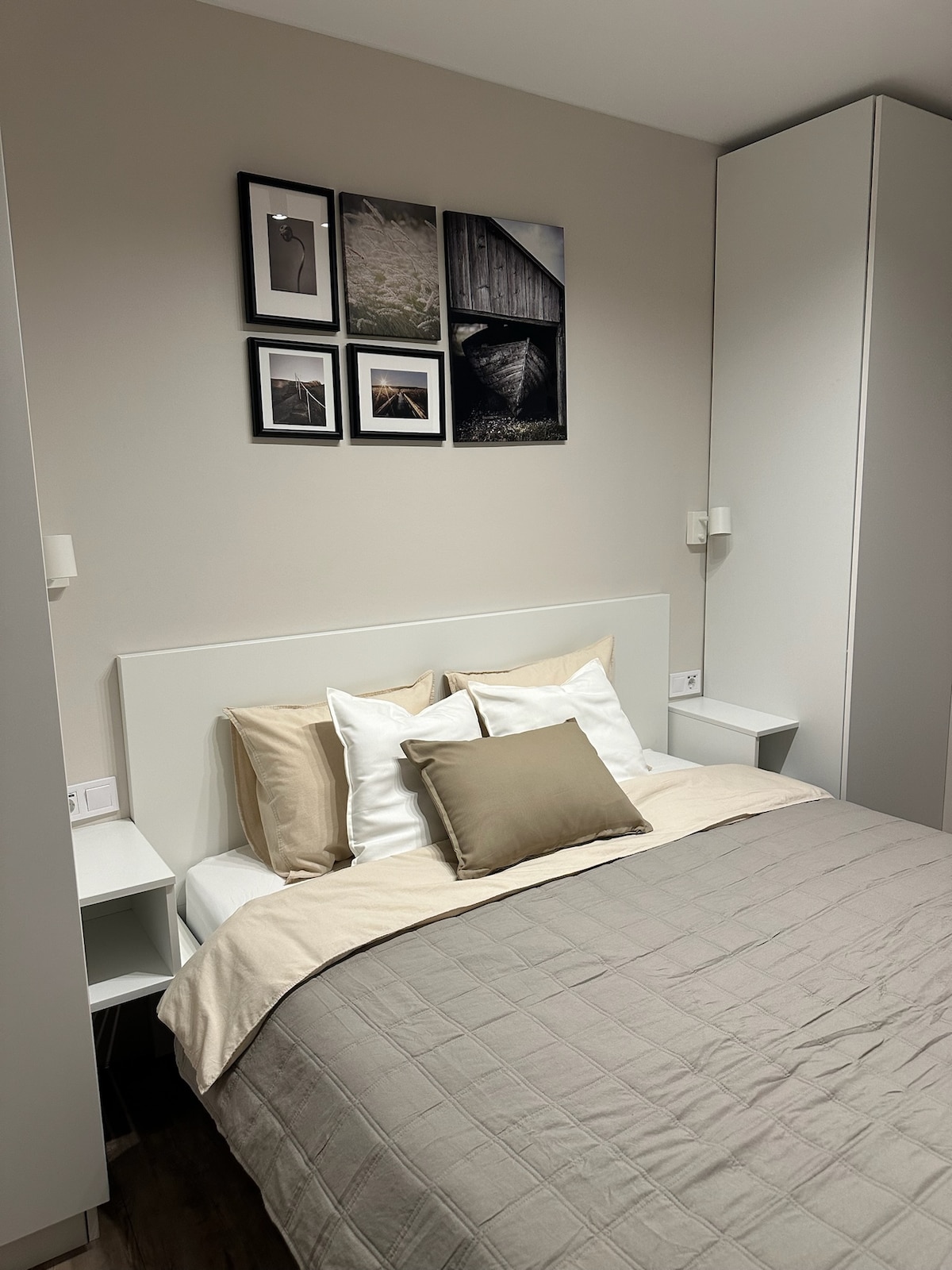 A serene bedroom features a spacious bed with layered bedding in neutral tones. Framed black and white photographs are displayed above the bed, complemented by sleek bedside tables. A built-in wardrobe is positioned to one side, enhancing the modern, minimalist aesthetic.