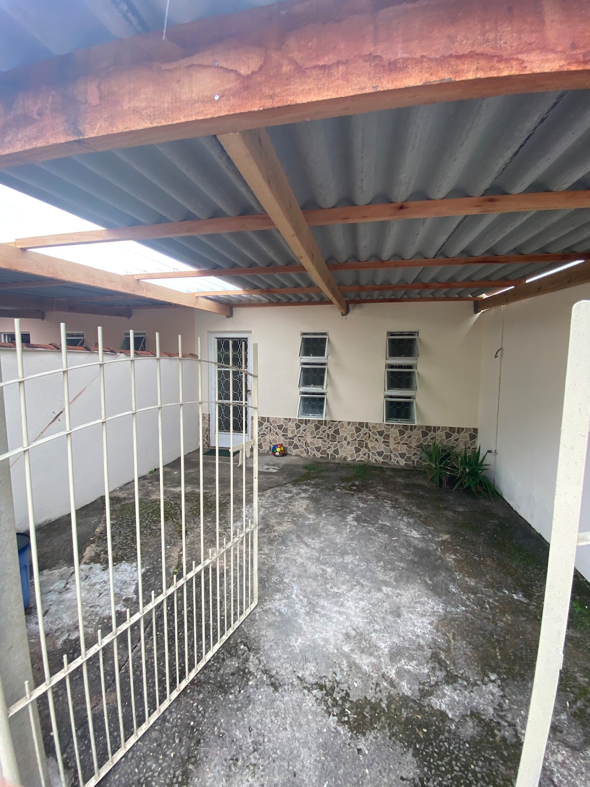 A gated outdoor area is presented, featuring a gravel surface with patches of greenery. The walls are smooth, and three narrow windows are visible, allowing light to enter. A wooden roof provides overhead coverage, ensuring a shaded space for relaxation.