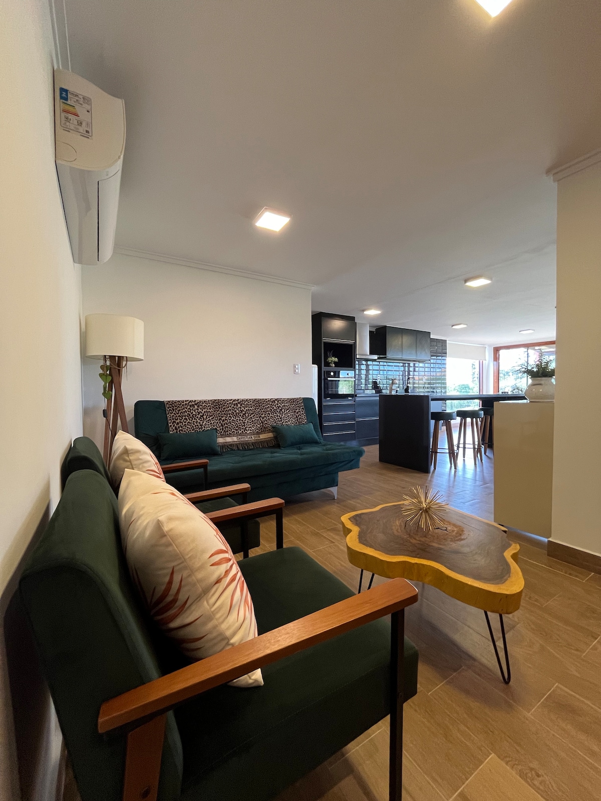 A stylish living area features a green velvet sofa and an armchair with decorative pillows. A small wooden table is placed between the seating. The open concept design allows for a view of the modern kitchen area in the background.