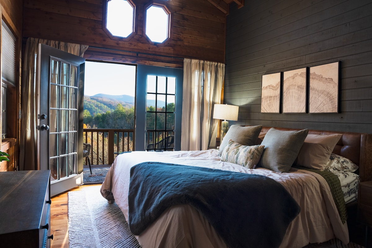 A spacious bedroom is accented by wooden walls and large windows offering mountain views. The king bed features plush pillows and blankets, with a cozy area rug beneath. French doors open to a private balcony, inviting natural light into the serene space.