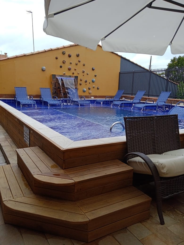 A swimming pool is framed by wooden decking, featuring several blue lounge chairs arranged around it. A gentle waterfall spills into the pool from a stone wall, while a large umbrella offers shade over a nearby seating area.