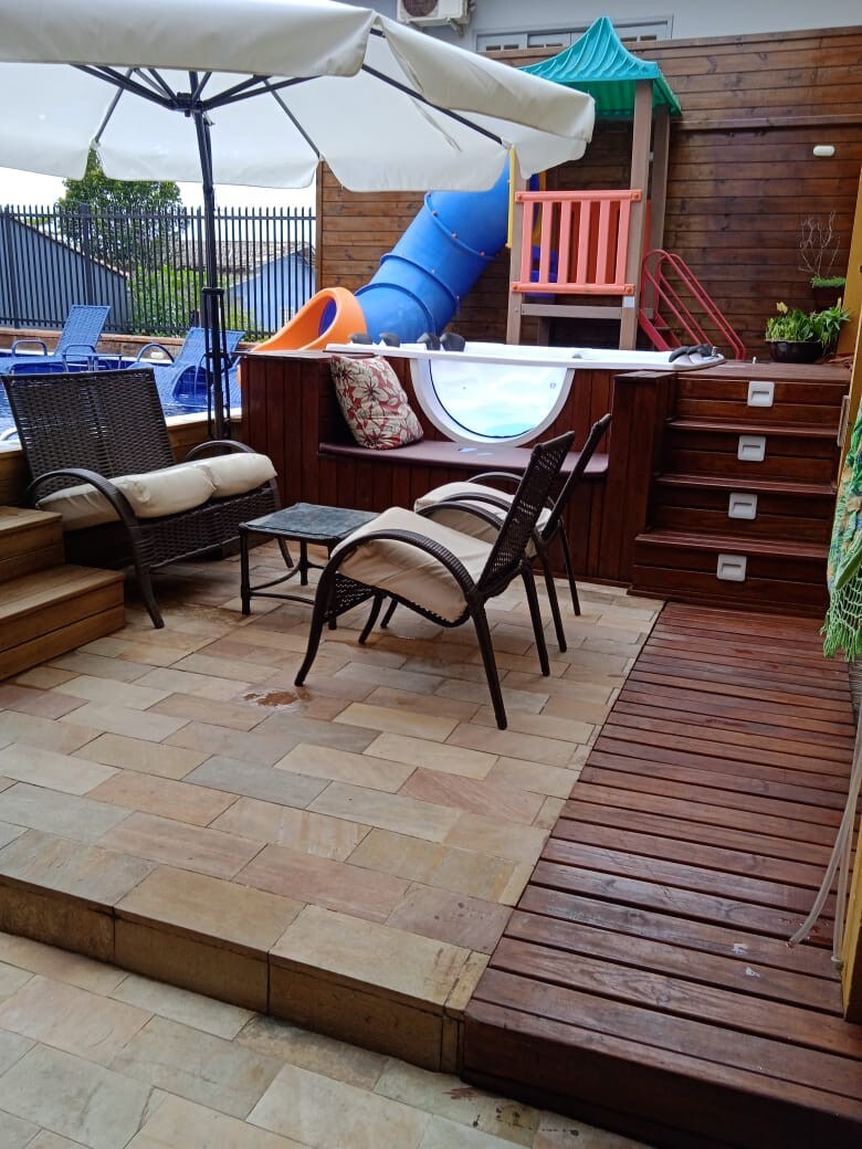 A recreational area is depicted with a seating arrangement consisting of two chairs and a small table. An umbrella provides shade, while a colorful water slide and play structure are visible in the background, enhancing the space for fun and relaxation.
