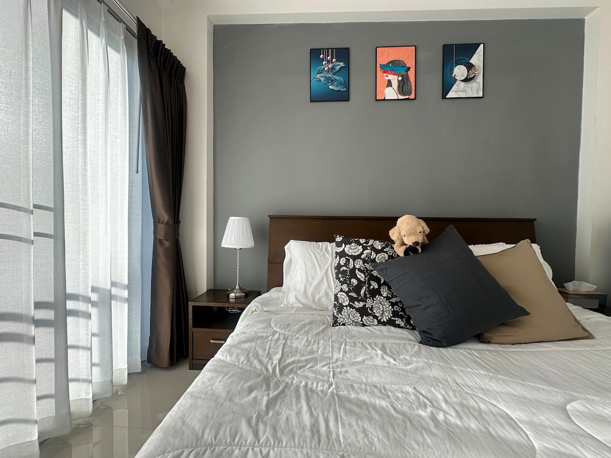 A private bedroom is presented with a spacious bed adorned with decorative pillows. A bedside lamp stands on a nightstand, and three framed artworks are displayed above the bed. Natural light is filtered through sheer curtains, complemented by dark drapes.