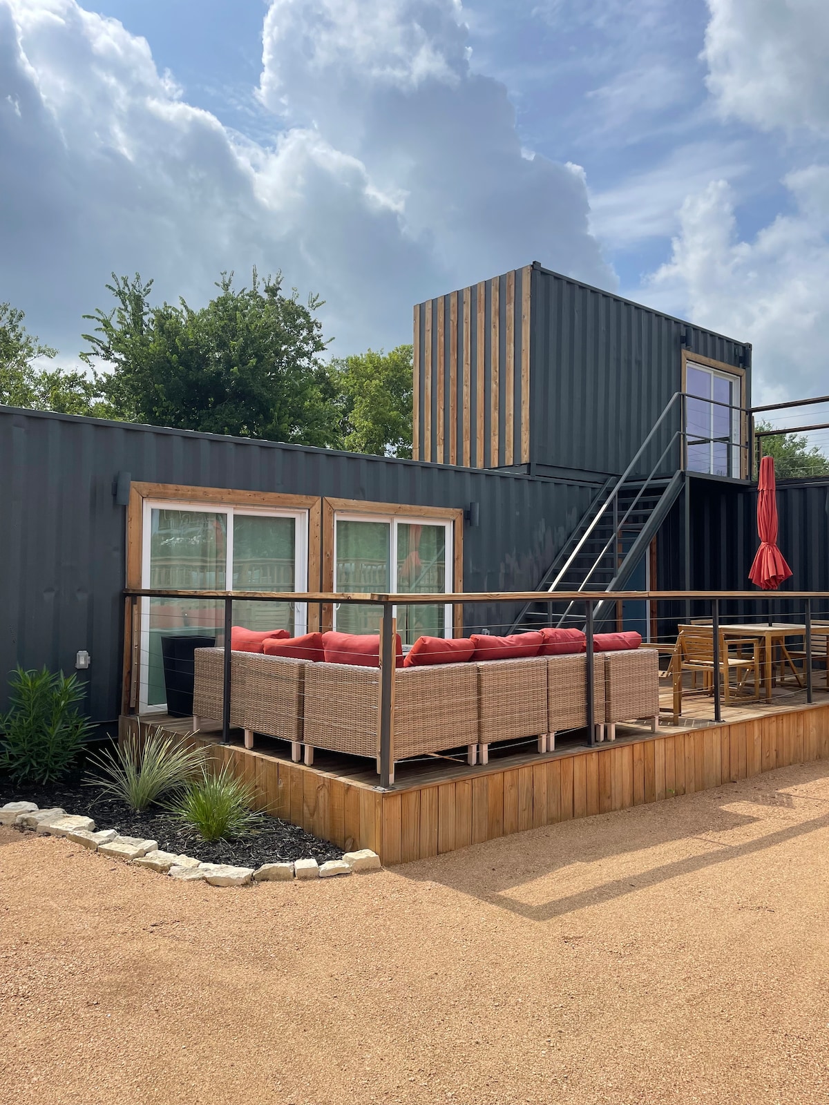 An outdoor seating area features a stylish arrangement of cozy couches with red cushions, surrounded by potted plants. The container structure, topped with a staircase leading to a second level, highlights large windows that bring in natural light. A vibrant red umbrella provides shade.