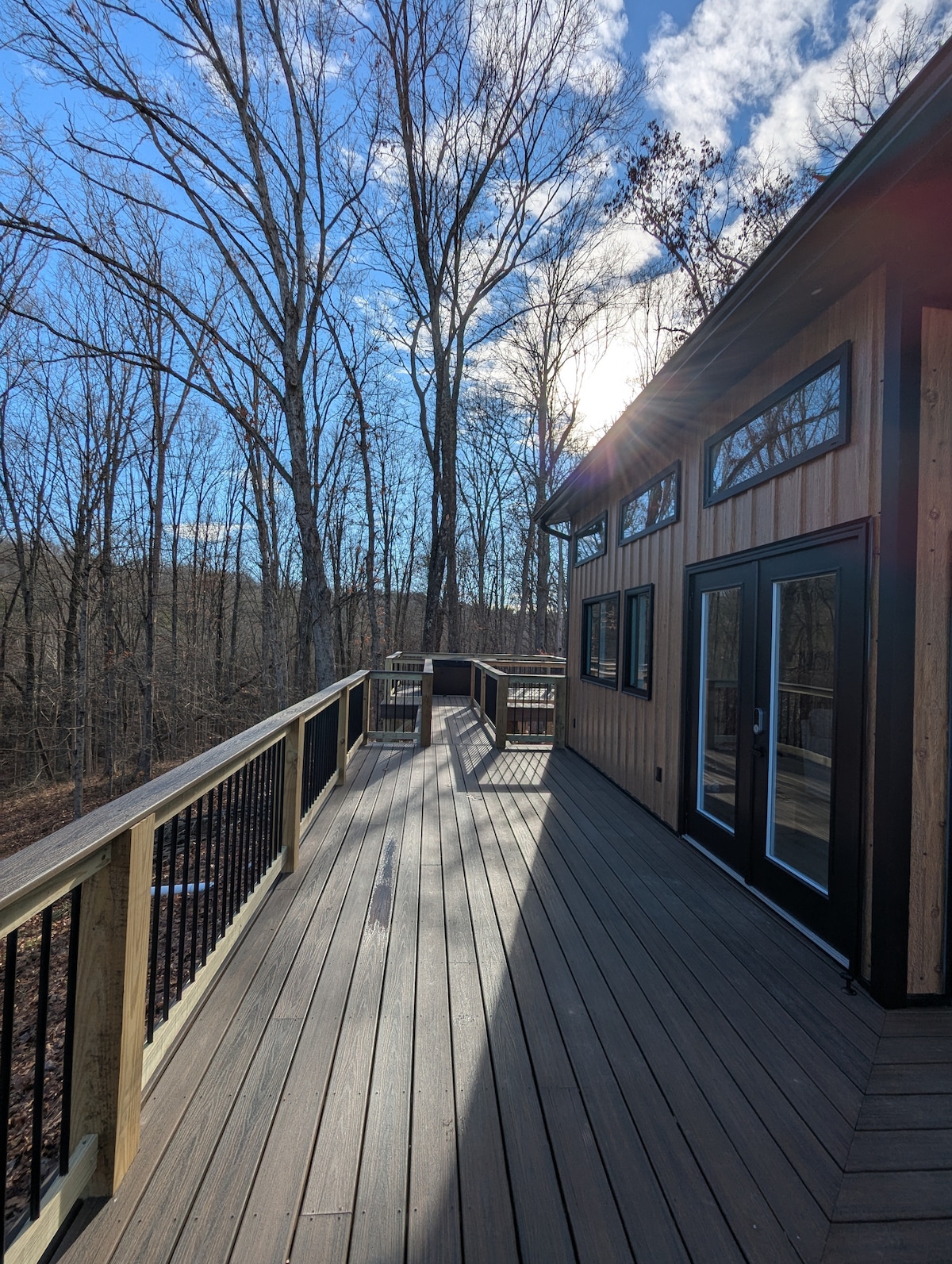 A spacious deck extends from the cabin, featuring wooden flooring and black railings. Sunlight filters through the trees, casting shadows on the deck. Large glass doors marked by black frames lead into the cabin, enhancing the connection with nature.