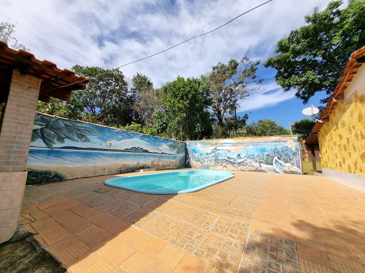 A refreshing swimming pool is set within a tiled area, surrounded by lush greenery and a vibrant mural depicting a seaside scene. The pool's clear water reflects the sky, creating a serene atmosphere, while the textured tiles offer a contrast to the natural elements.